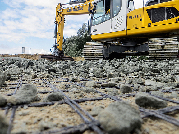 Base course reinforcement with geogrids An excavator distributes bulk material on the installed geogrid